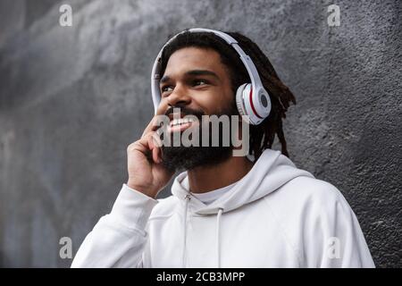Picture of a positive cheery young man walking outdoors while listening music with headphones