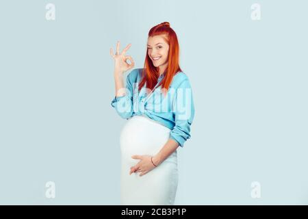 Content redhead girl in expectancy showing OK gesture and winking at camera happily isolated on blue background. Everything is alright with my pregnan Stock Photo