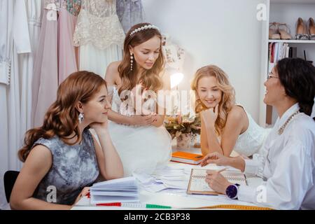 cheerful smiling group of young caucasian and beautiful models gathered in salon to listen to, get consultation and try on new wedding dresses made by Stock Photo
