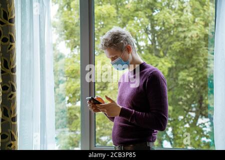 Young caucasian man wearing face mask while using his mobile phone standing near window. Lifestyle photo with copy space. New normal rules for staying Stock Photo