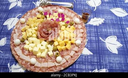 White dish decorated with salami, ham, quail eggs and heart of palm, on a background with a blue and white tablecloth, with a whiskey glass handle Stock Photo