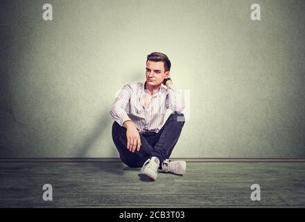 Portrait of thoughtful young man in stripped white light shirt thinking sitting on grey floor in front of gray background in studio. Horizontal shot m Stock Photo