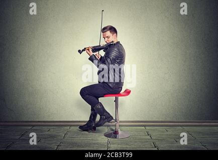 Music passion, hobby concept. Young man dressed modern and elegantly in black leather jacket playing on black violin sitting on red chair in studio is Stock Photo