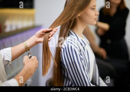 side view on beautiful caucasian girl sitting on chair in beauty salon, closeup of long hair, hairdresser hands holding them, going to cut split ends. Stock Photo