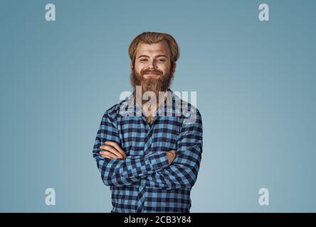 Portrait of young confident smiling man. Hipster male with beard in blue plaid checkered shirt  Isolated on blue studio Background. Negative face expr Stock Photo