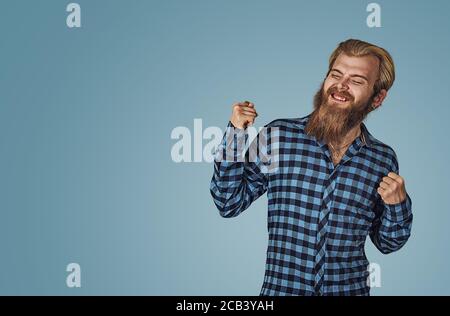 Happy successful student man winning, fists pumped celebrating success. Hipster male with beard in blue plaid checkered shirt  Isolated on blue studio Stock Photo