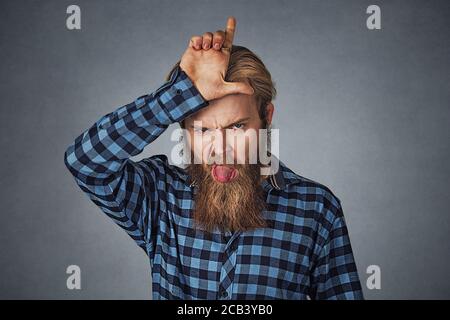 Portrait of a funny young bearded man holding finger up at his forehead and sticking tongue out. Hipster male with beard in blue plaid checkered shirt Stock Photo