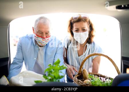 Senior couple with face masks putting shopping in car outside supermarket in city. Stock Photo