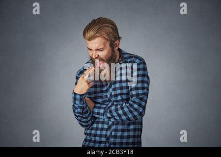Disgusted man with finger in mouth displeased wants to throw up. Hipster male with beard in blue plaid checkered shirt  Isolated on gray grey studio B Stock Photo