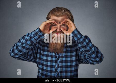 Displeased young man looking through fingers like binoculars. Hipster male with beard in blue plaid checkered shirt  Isolated on gray grey studio Back Stock Photo