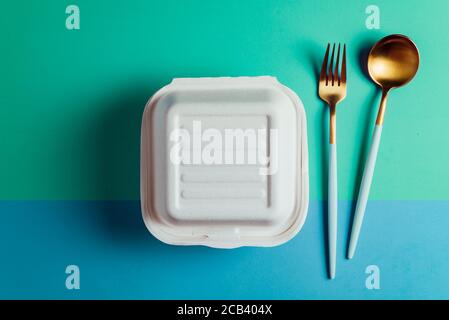 Healthy food concept: white burguer packaging closed with golden fork and spoon,  in cardboard lunch box to take to the office on blue and green surfa Stock Photo