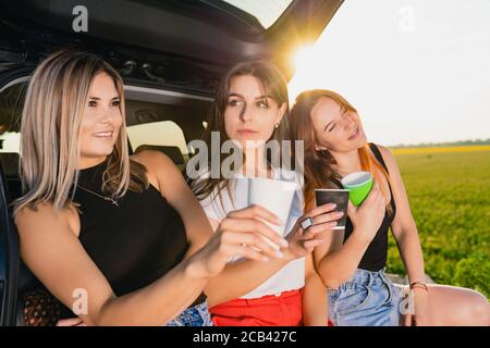 Happy road trip of three smiling female travelers, relax at vehicle back with coffee cups take away at sunlight. Stock Photo