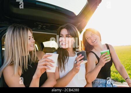 Happy road trip of three smiling female travelers, relax at vehicle back with coffee cups take away at sunlight. The girls are drinking coffee. Stock Photo