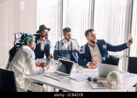 Positive caucasian bearded businessman taking selfie with his middle eastern and indian partners during workday, sitting in conference room. Stock Photo