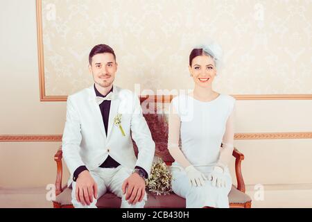 Content elegant man and woman in wedding outfits sitting on sofa in ornamental interior looking at camera Stock Photo