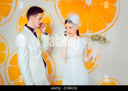 Trendy man and stressed woman in wedding outfits having phone calls while standing against wall with oranges Stock Photo