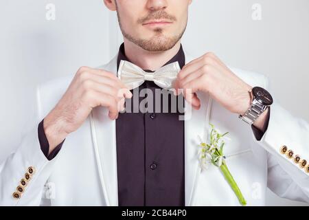 Crop shot of young groom in classy white suit with black shirt adjusting white bow tie Stock Photo