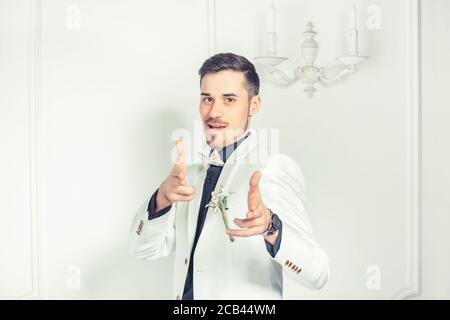 Trendy young man in groom suit pointing at camera with excitement hands shaped like guns telling is your turn now on white classical background Stock Photo