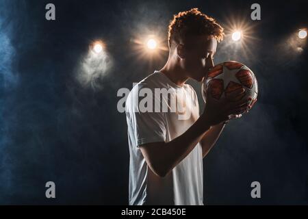 Young adult football player kissing football ball against black background Stock Photo