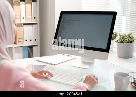 Over shoulder view of Muslim programmer sitting at office table and coding information for website using computer Stock Photo