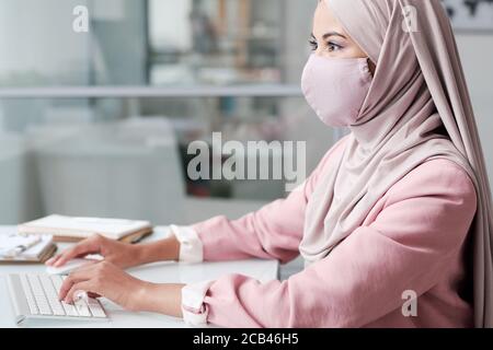 Side view of busy young Muslim woman in facial mask working in office Stock Photo