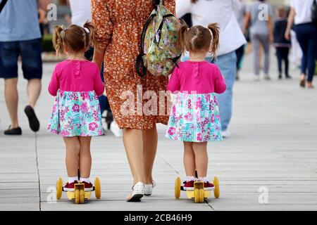 Mom and twin kids on a scooters are walking the street. Young slender woman lead little daughters in identical clothes, concept for motherhood Stock Photo