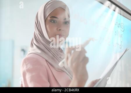 Behind glass board view of concentrated young Arabian programmer in hijab writing data while analyzing code Stock Photo