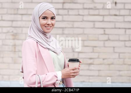 Portrait of positive beautiful young Muslim woman in hijab standing against brick wall and drinking coffee on move Stock Photo