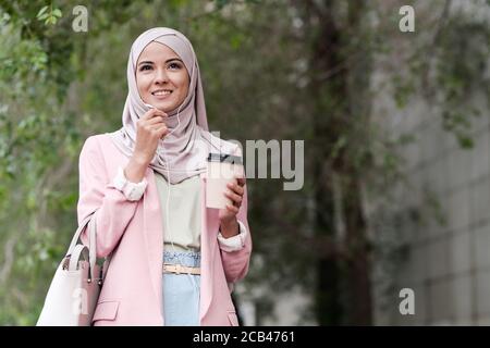 Positive confident young Muslim businesswoman in stylish outfit drinking takeaway coffee and talking through phone headset outdoors Stock Photo