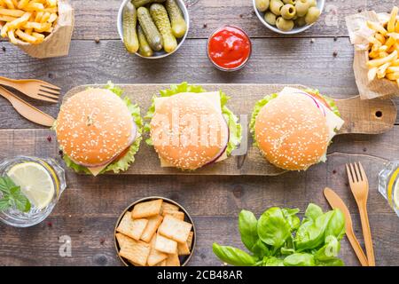 Dinner table with burger, french fries, vegetables, sauces, snacks and lemonade on wooden background, top view. Stock Photo
