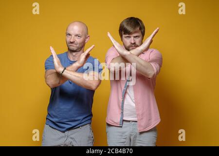Two serious, confident men in casual clothes shows gesture closed path with arms crossed, on a yellow background. Stock Photo