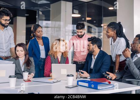 Creative multiracial business team discussing project, standing around workplace, while two co-workers with portable computers looking for needed info Stock Photo