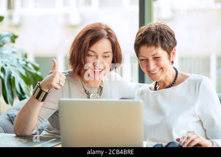 Contemporary aged women in trendy clothes having online call while using laptop and showing thumb up Stock Photo
