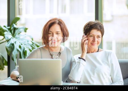 Elegant woman using laptop while sitting with friend speaking on phone at table in cafe Stock Photo