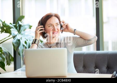 Modern middle-aged woman wearing headset and sitting at table with laptop smiling away Stock Photo