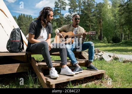 Mixed race guy sitting on porch of tent and listening how black friend playing guitar Stock Photo