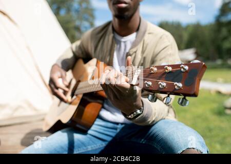 Close-up of young Black man sitting on wooden porch of camping tent and playing guitar outdoors Stock Photo