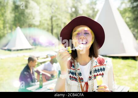 Portrait of happy attractive girl in hat making soap bubbles through bubble stick at campsite Stock Photo