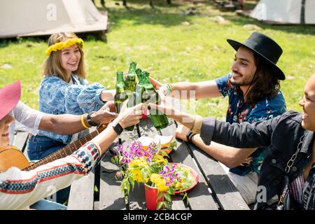 Group of cheerful young multi-ethnic friends sitting at outdoor table and toasting with bottles at campsite Stock Photo