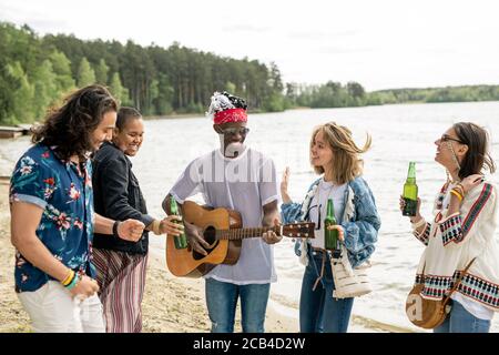 Group of positive young multi-ethnic friends dancing on beach under guitar music by hippie black guy Stock Photo