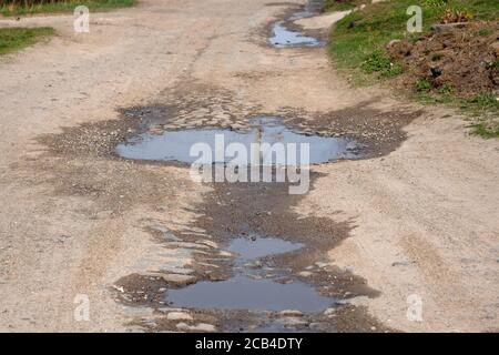 Broken country road with pits in the spring. The road without asphalt with deep puddles. A large puddle on a dirt road along a street in a village. Stock Photo