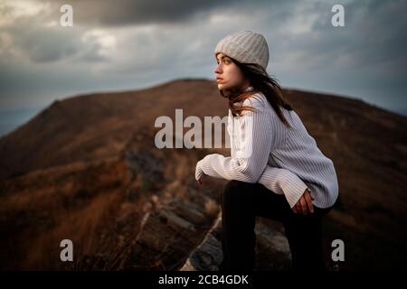 Young beautiful woman in a white sweater stands on top of a mountain and enjoys the scenery. Young woman standing on cliff's edge and looking to a sky Stock Photo