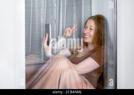 happy smile girl sits on a windowsill by the window and holding a smartphone in one hand with the other hand shows the gesture of peace, working remot Stock Photo