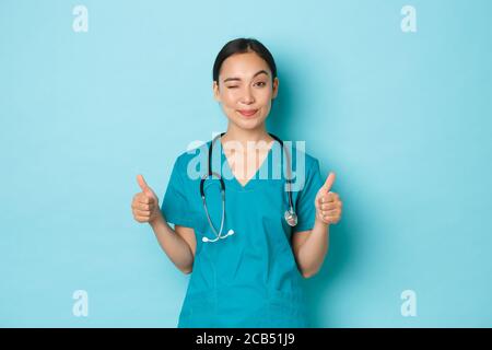 Covid-19, social distancing and coronavirus pandemic concept. Sassy beautiful asian girl in scrubs encourage visit her clinic, winking and showing Stock Photo