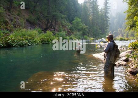 Human fly fishing in deep blue pond in river with guide standing on the edge of the water with scooping net and provide guidance Stock Photo