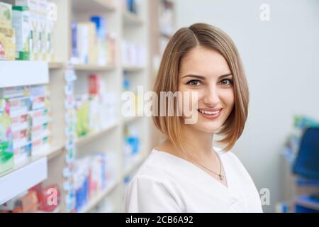 Portrait of smiling female pharmacist standing near shelves with medicaments in drugstore. Side view of woman in white lab coat looking at camera and posing in pharmaceutical store. Medicine concept. Stock Photo