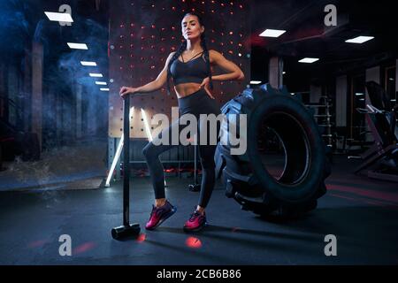 Muscular woman holding big sledgehammer near huge tire wheel, looking away. Portrait of female bodybuilder posing in gym, having rest after hard training and looking at camera. Concept of sport. Stock Photo