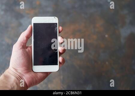 A young man holds a smartphone with a broken screen in his hand on a dark background. Mockup Stock Photo