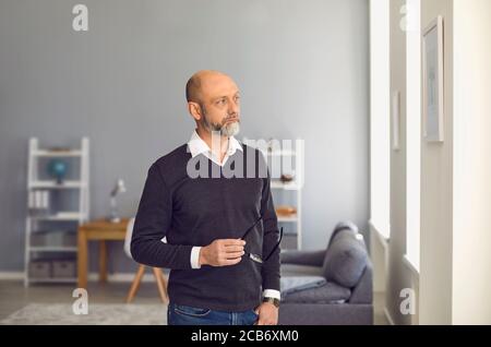 Serious man senier with glasses standing looking away while standing in a room at home. Stock Photo