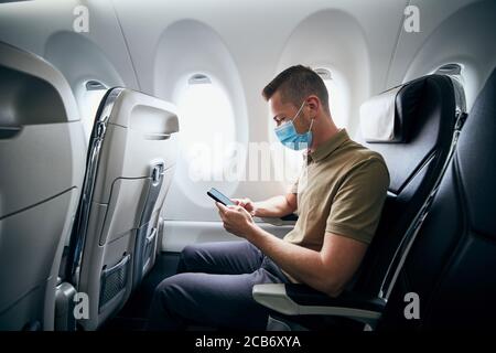 Man wearing face mask and using phone inside airplane during flight. Themes new normal, coronavirus and personal protection. Stock Photo